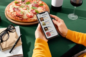 A person holding a smartphone browses a restaurant listing app over a green tablecloth. Next to the phone on the table sits a freshly baked pizza, a glass of red wine, a wine bottle, glasses, and a notebook.