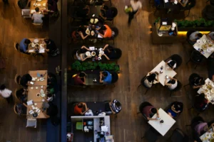 An overhead view shows patrons seated at various tables inside a busy restaurant with wooden flooring. People are eating, chatting, and using their smartphones while staff move between the dining areas.