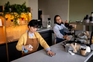 Two café workers wearing aprons clean the counter area inside a modern coffee shop. In the foreground, a woman sprays and wipes down the countertop while a man behind her cleans the espresso machine.