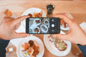 A top-down view shows a person holding a smartphone to take a photo of a meal spread across a wooden table. The dining table features plates with burgers, french fries, other dishes, and drinks.