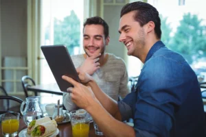 Two smiling young men sit together at a café table while looking at content on a digital tablet held by the man in the blue shirt. Food, orange juice, and water are set on the table in front of them near a brightly lit window.