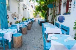 A cobblestone alleyway in a Mediterranean-style village is lined with whitewashed buildings and outdoor restaurant seating. Tables are set with white and blue-dotted tablecloths alongside bright blue chairs, wooden stump stools, and lush green foliage overhead.