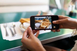 A person holds a smartphone to capture a photo of a diner meal at a green table. The phone screen clearly shows toasted sandwiches served with French fries and a side sauce.