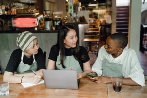 Three cafe workers wearing matching green aprons sit around a wooden table inside a coffee shop, collaborating with an open laptop. One employee takes notes on a notepad while another holds a smartphone as they chat together.