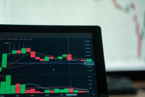 A close-up shot shows a laptop screen displaying a dark-mode financial trading chart with red and green candlestick patterns and trend lines. In the blurred background, another display reflects additional stock or cryptocurrency market graphs.