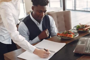 A man wearing a vest sits at a dining table with salads and a laptop nearby, looking down thoughtfully. Next to him, a person in a white button-up shirt leans over to write notes in a spiral notebook on the table.