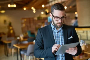 A bearded man wearing glasses and a dark blazer focuses intently on a digital tablet in his hands. He is standing in a warmly lit, modern restaurant or cafe setting.