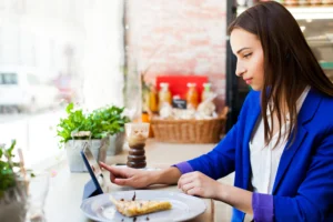 A woman wearing a bright blue blazer sits at a window counter in a bright cafe, focused on using a digital tablet. On the wooden counter beside her sit a slice of pie on a plate, an iced drink, and small potted plants.