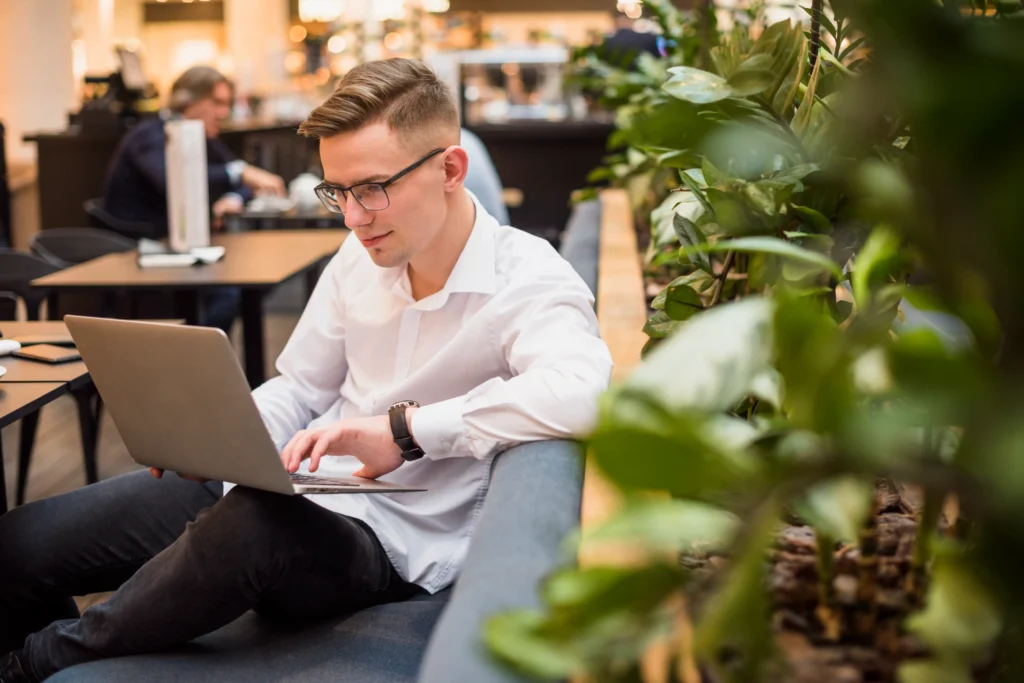A young man wearing glasses and a white button-up shirt sits on a sofa, focused on typing on his laptop. He is working in a modern, well-lit cafe or lounge space lined with indoor plants.