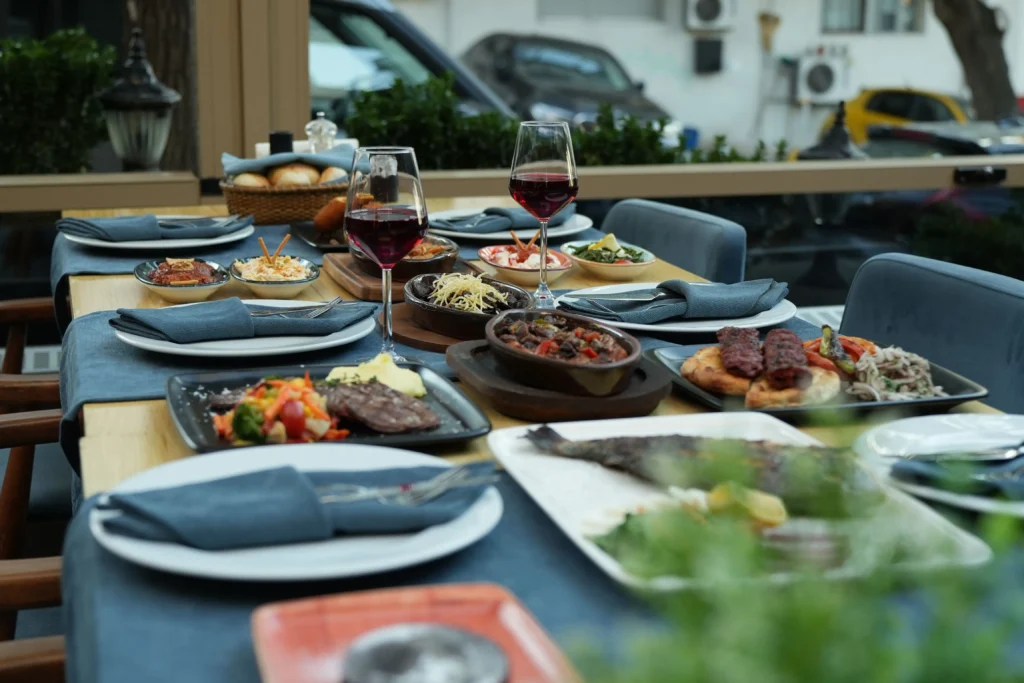 An outdoor restaurant table is abundantly set with various Mediterranean dishes, including grilled meats, fresh fish, stews, and small appetizers. Glasses of red wine sit alongside blue cloth napkins and full place settings for an al fresco dining experience.