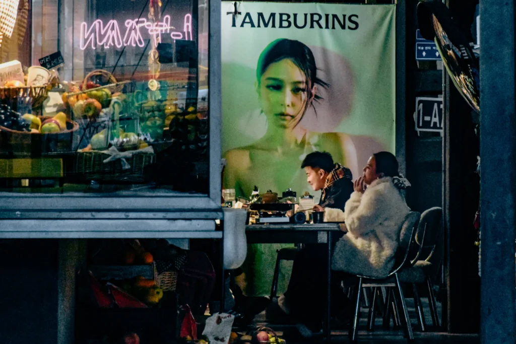 Two people sit at an outdoor dining table at night, eating in front of a large poster for the brand Tamburins featuring Blackpink's Jennie. Beside them, a lit store display case filled with fresh fruit adds warm ambient light to the street scene.