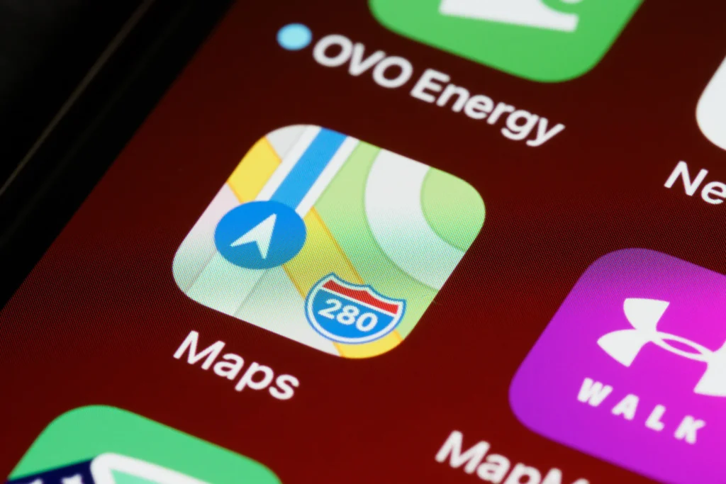 A close-up shot displays the Apple Maps app icon centered on a smartphone screen. Surrounding apps including OVO Energy and Under Armour MapMyWalk are partially visible against the dark wallpaper.