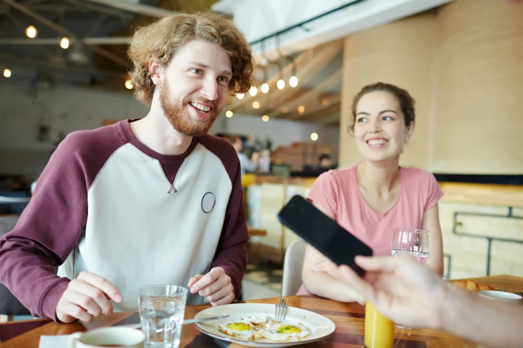A man with red hair and a woman sit at a table in a brightly lit restaurant while enjoying breakfast and drinks. They both smile as someone out of frame holds up a smartphone toward them.