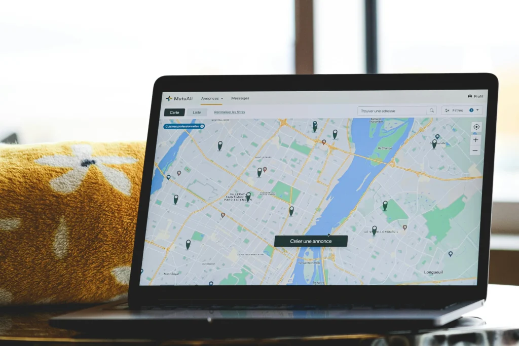 An open laptop rests on a surface, displaying an interactive map web application with several location pins across a city area. Beside the device, a cozy yellow floral-patterned blanket adds a warm accent to the brightly lit indoor setting.