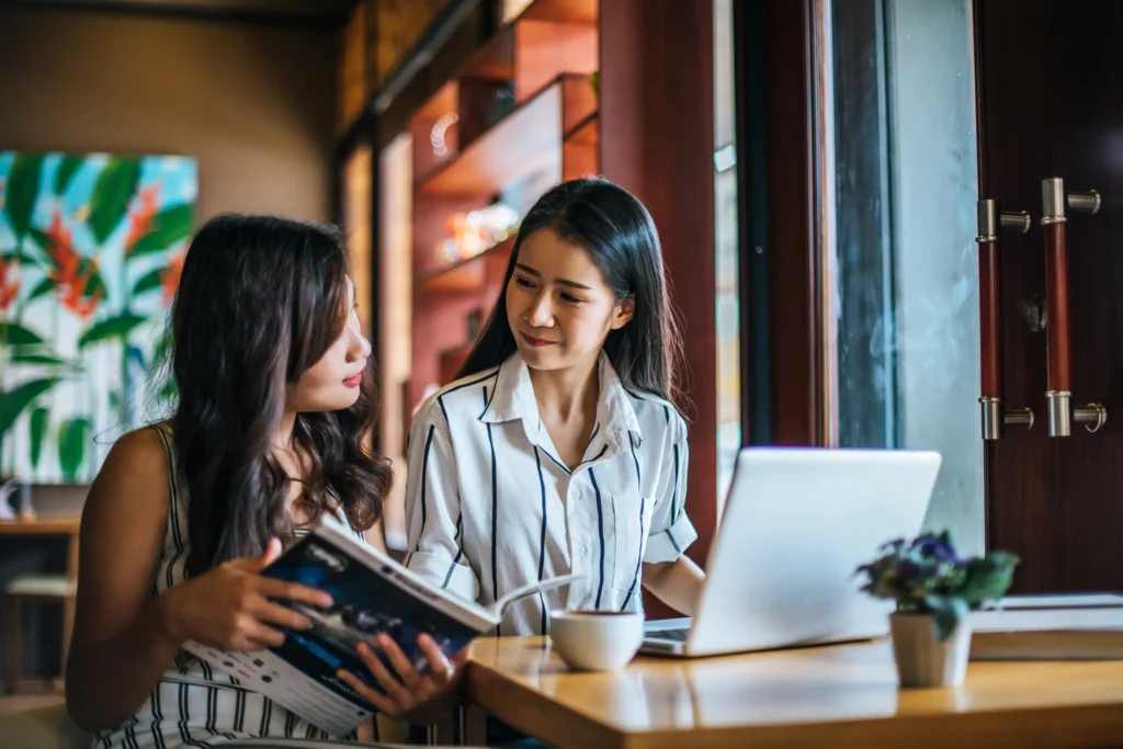 Two young women sit together at a wooden table inside a cozy cafe near a window. One woman holds open a magazine while looking toward her friend, who sits in front of an open laptop with a cup of coffee on the table.
