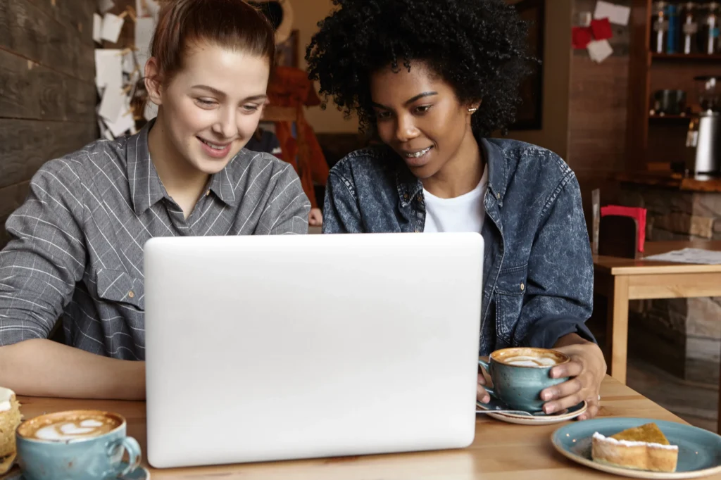 A man in a plaid shirt and a woman in a red top sit together at a dark table in a cozy café or restaurant, both focused on an open laptop. They hold small white coffee cups as they intently view the screen, with a flower vase and candle decorating the table beside them.