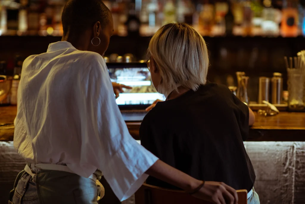 Two women sit side by side at a warmly lit bar counter, looking intently at an open laptop screen together. One woman, wearing a white shirt and apron, points toward the screen while resting her hand on the back of the other woman's chair.