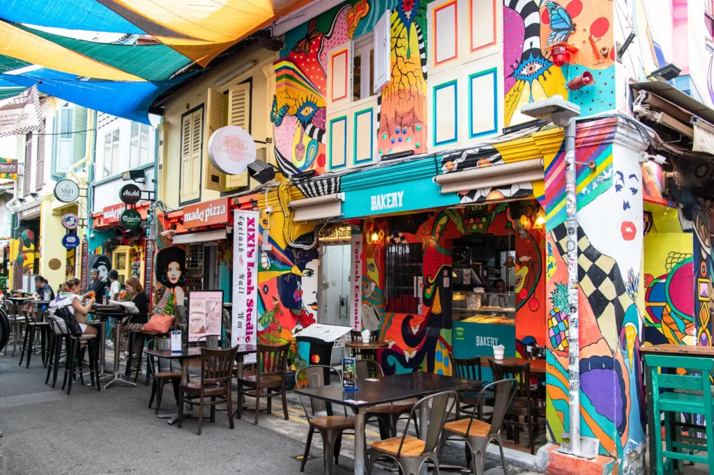 A vibrant street scene features historic storefronts covered in colorful, psychedelic murals with outdoor dining tables set along the pavement. Multi-colored sunshades stretch overhead across the narrow alleyway, creating a lively atmosphere for nearby cafes and shops.