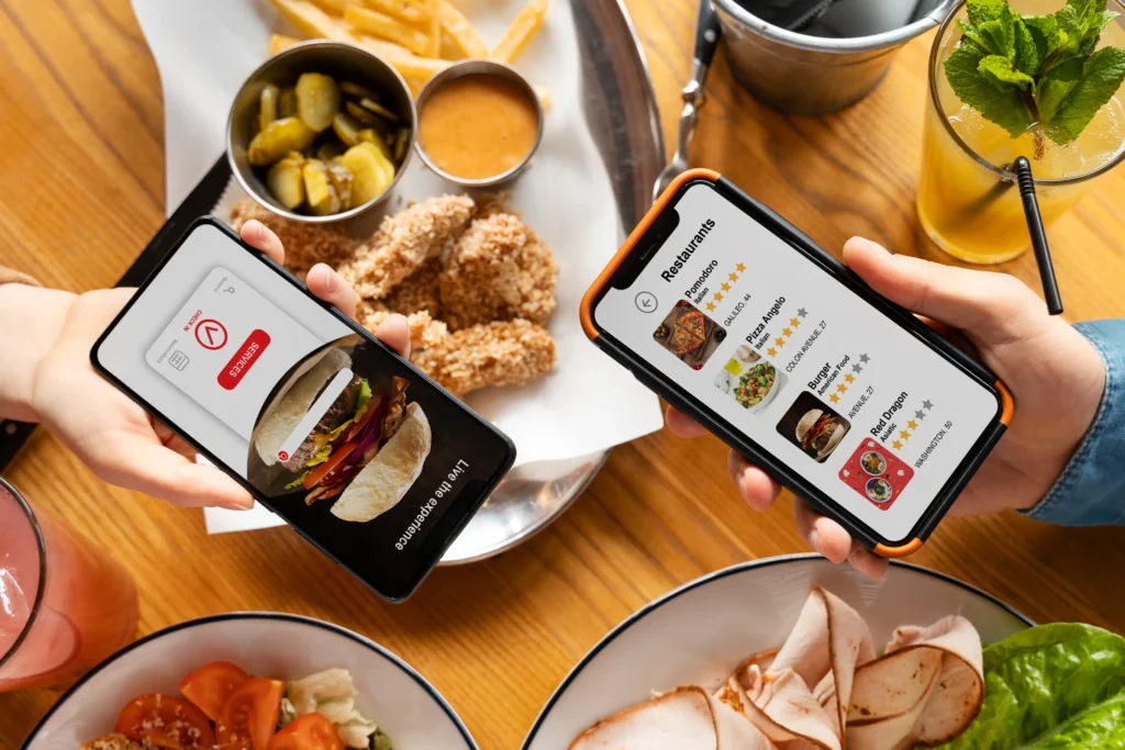 Two people hold smartphones over a dining table filled with dishes including fried chicken tenders, french fries, and salads. One screen displays a food service application, while the other shows a list of restaurant recommendations with star ratings.