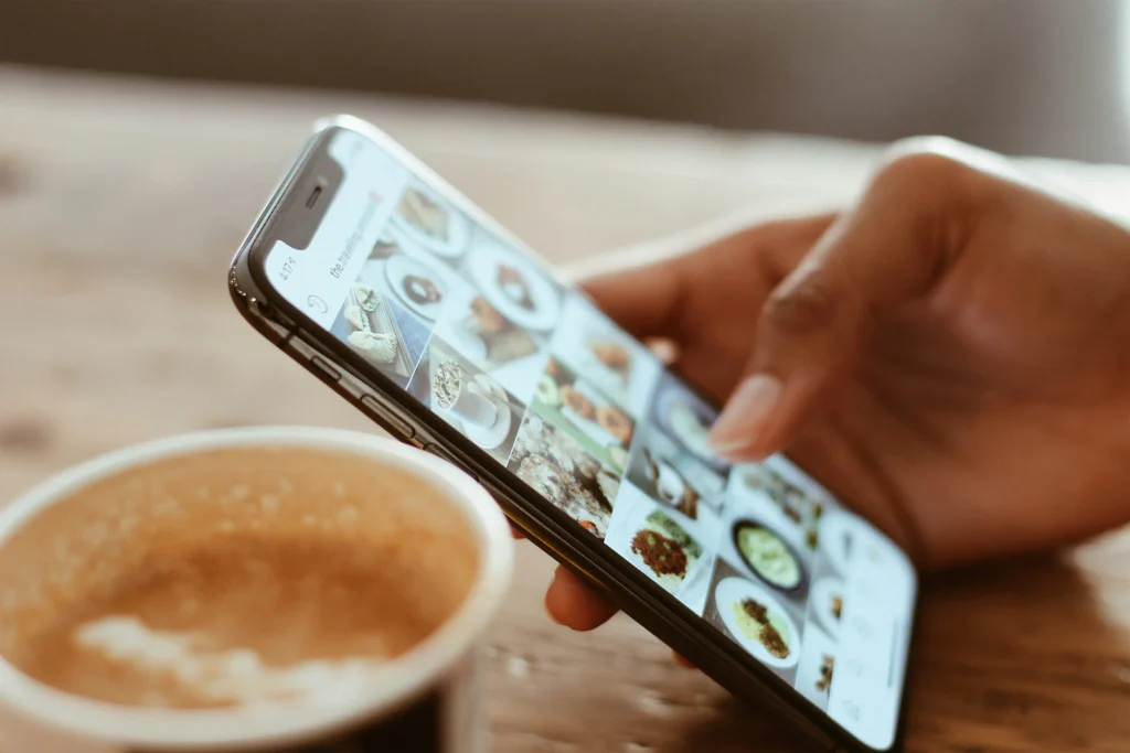 A close-up shot of a person holding a smartphone and scrolling through a visual grid feed, likely Instagram. In the soft-focus foreground sits a cup of hot coffee on a wooden table.
