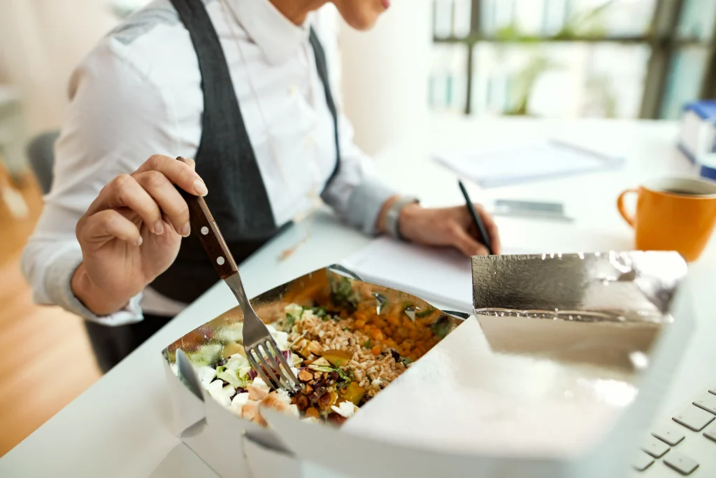 A person wearing a white collared shirt and vest uses a fork to eat a healthy salad bowl from a foil takeout container. They are sitting at a office desk and writing on a notepad with a pen in their other hand while taking a lunch break.