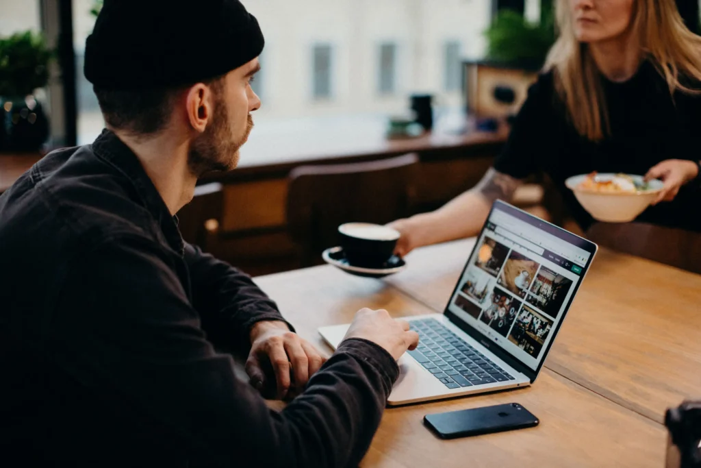 A man wearing a black beanie sits at a wooden table in a cafe, working on an open laptop. Across from him, a woman hands over a cup of coffee while holding a bowl of food.