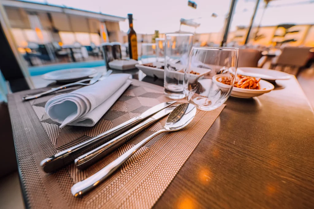 A close-up shot of an elegant restaurant table set with shiny polished silverware, drinking glasses, plates, and a neatly rolled white cloth napkin on a woven placemat. In the softly blurred background, warm lighting reflects off the contemporary dining space near a pool.