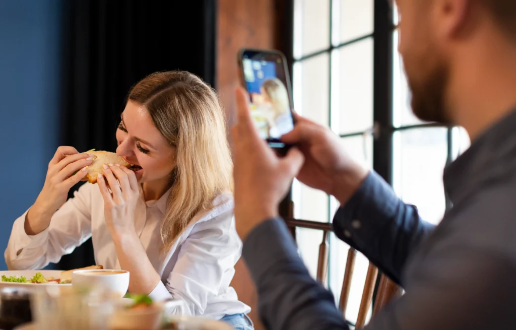 A smiling blonde woman takes a bite of a sandwich at a cozy restaurant table. In the foreground, a man holds up a smartphone to snap a photo of her.