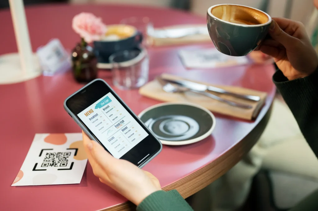 A high-angle view of a person seated at a café table holding a smartphone displaying a digital menu. In their other hand, they hold a cup of coffee above a table set with a printed QR code card, empty saucer, and cutlery.