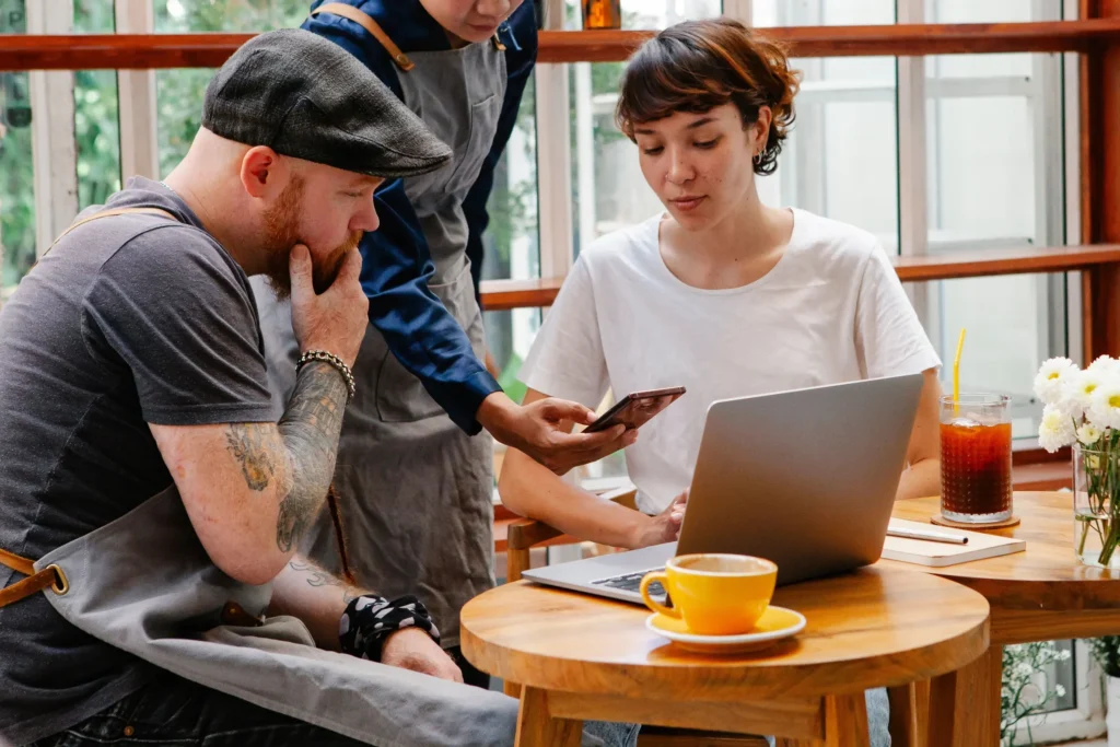 A person wearing an apron holds out a smartphone to show something on screen to a customer at a cafe table. The customer seated behind an open laptop looks attentively at the phone screen while another person sitting beside them looks on thoughtfully.