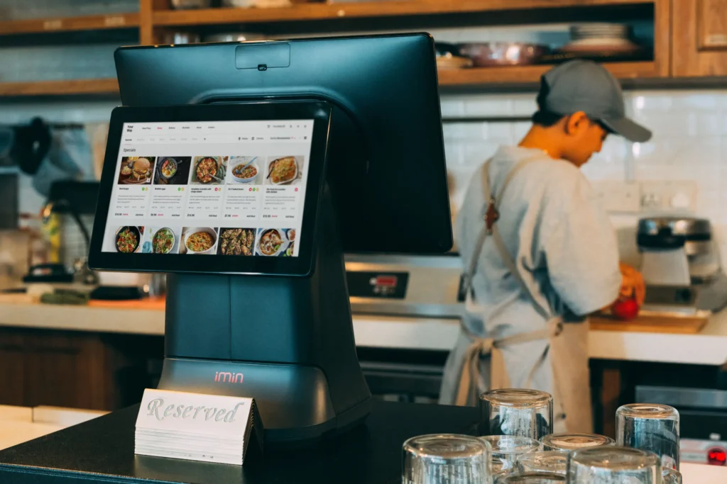 A digital POS register displaying a food ordering menu sits on a cafe counter next to a "Reserved" sign. In the background, a worker wearing a cap and apron prepares food in the kitchen area.
