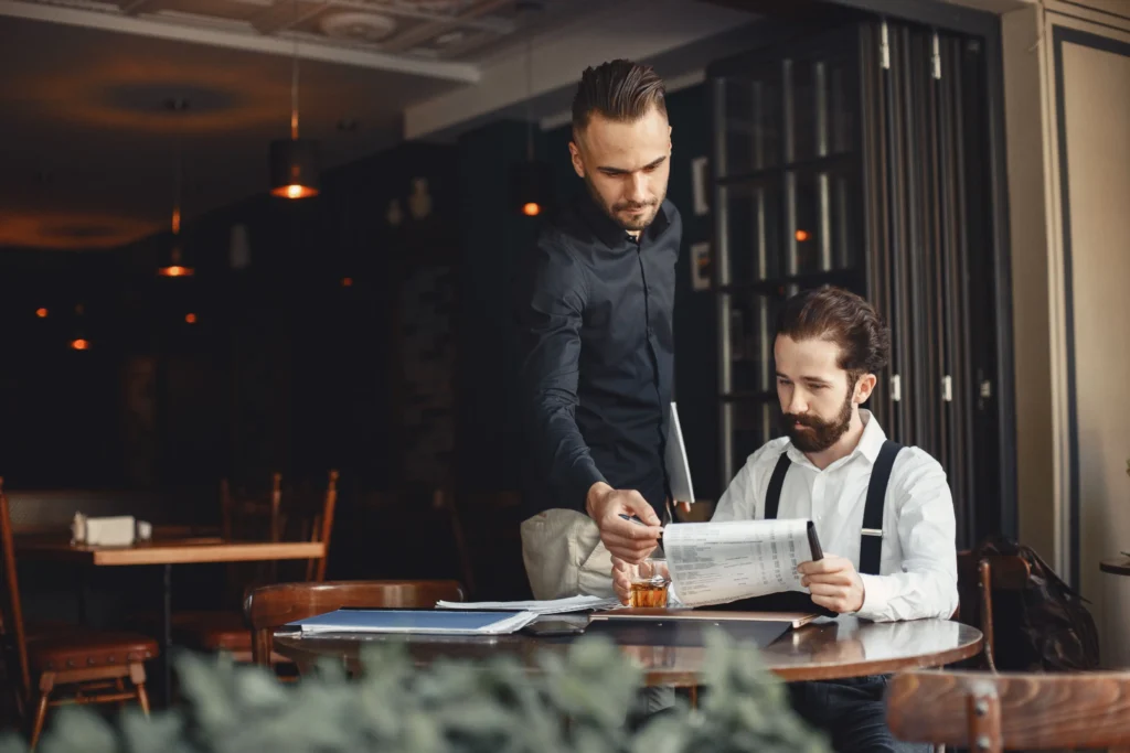 A man standing at a café table points to a document while holding a notebook, assisting a seated colleague with a full beard and suspenders. Together, they review paperwork and files spread across the round wooden table.