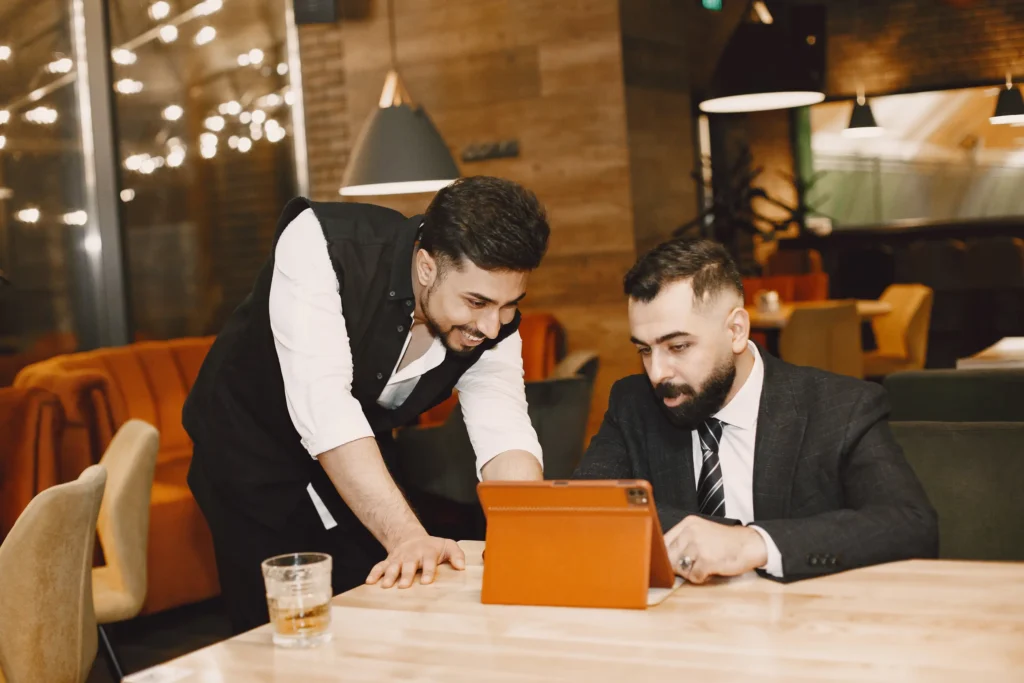 Two men in professional attire review content together on a tablet resting on a wooden table. One man sits in a suit jacket while the other leans over to look at the screen inside a dimly lit restaurant setting.