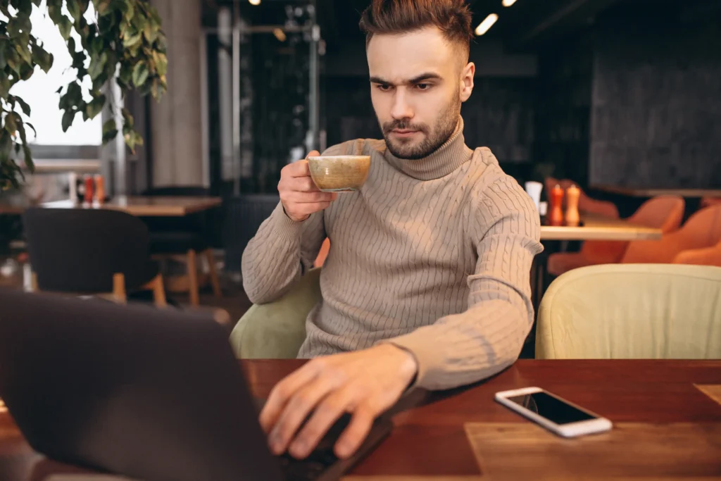A focused man in a beige turtleneck sweater sits at a wooden table in a cafe while typing on his laptop. He looks intently at the screen while holding a small cup of coffee in his other hand.