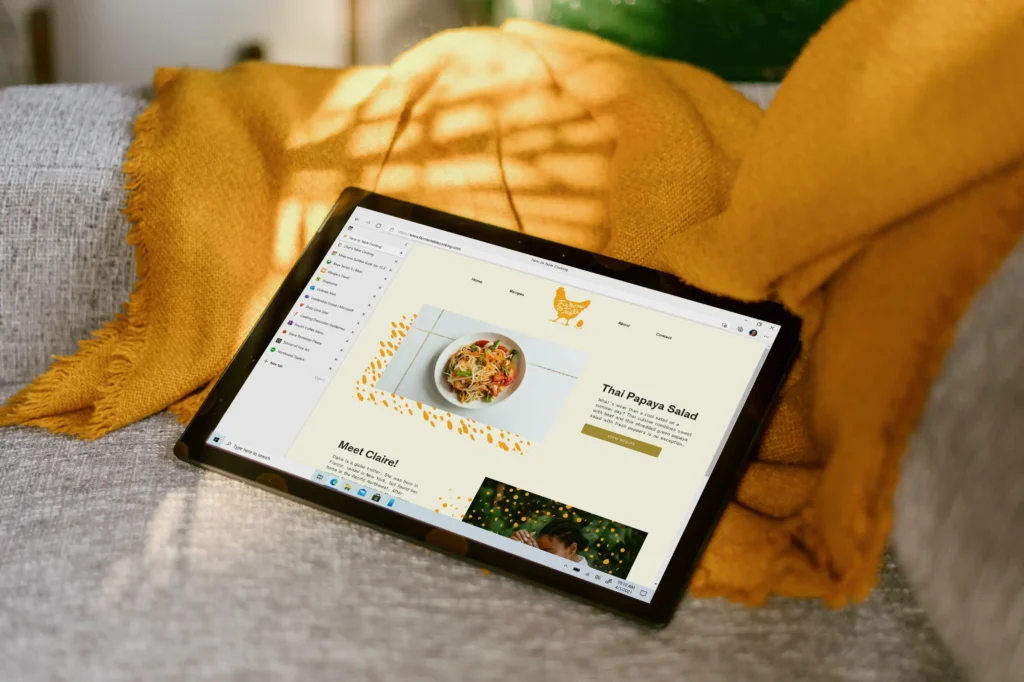 A tablet rests on a grey sofa beside a yellow fringed blanket under warm, patterned sunlight. The screen displays a food blog featuring a recipe for Thai Papaya Salad with a yellow chicken logo at the top.