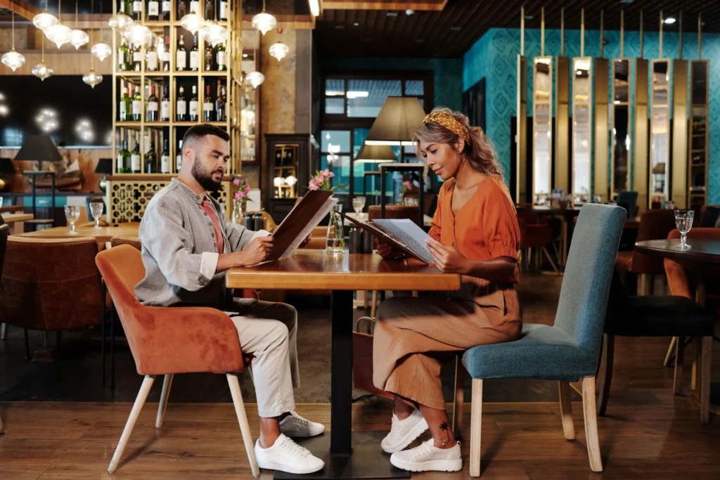 A man and a woman sit across from each other at a wooden table in an upscale, dimly lit restaurant while looking over open menus. The modern dining space features a tall wine rack, warm overhead lighting, and stylish contemporary decor.