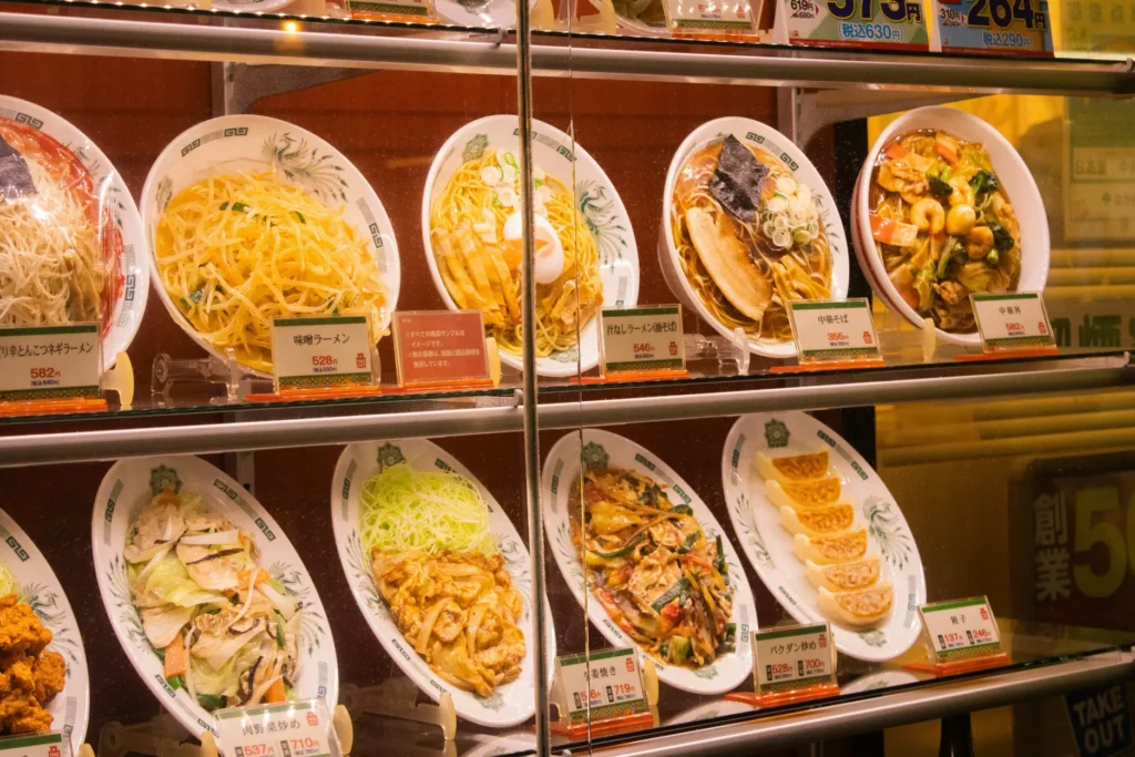 A glass display case outside a restaurant showcases realistic plastic food models of various Japanese noodle and stir-fry dishes. Each plate features a small price card underneath detailing the menu item and cost.
