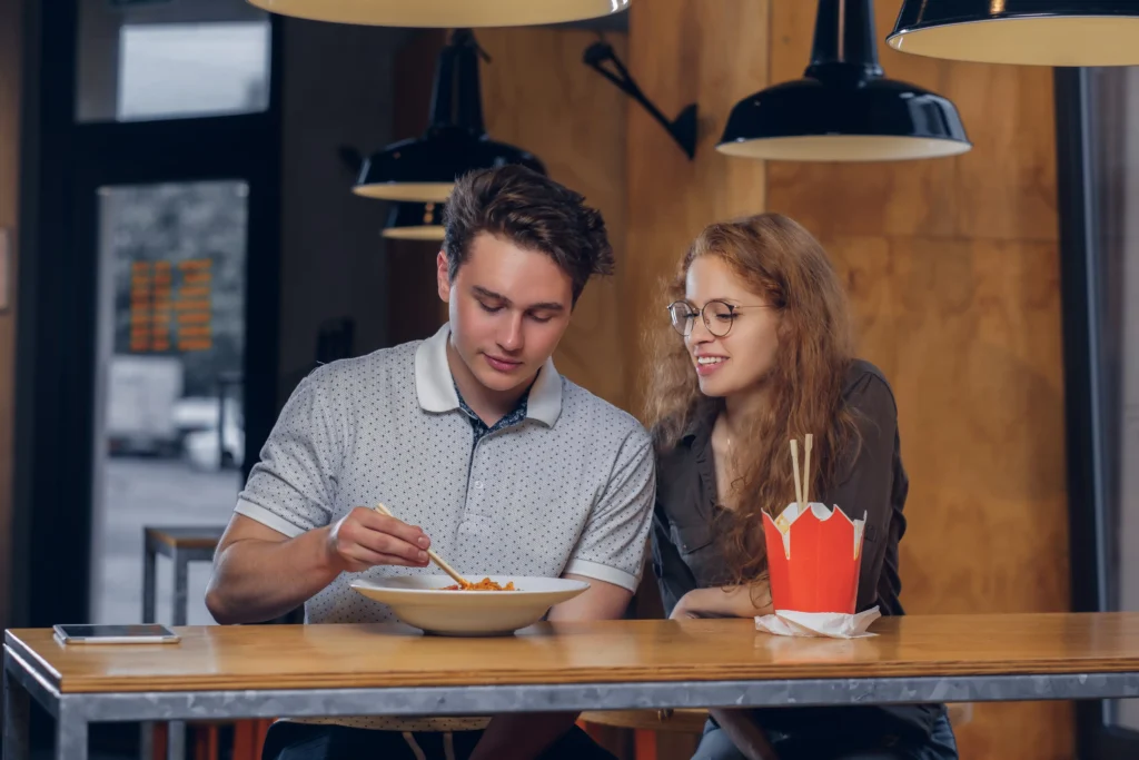 A young couple dressed in casual clothes sits together at a wooden counter in a restaurant. The man uses chopsticks to eat noodles from a bowl while the woman watches him with a smile, a red takeout box resting nearby.