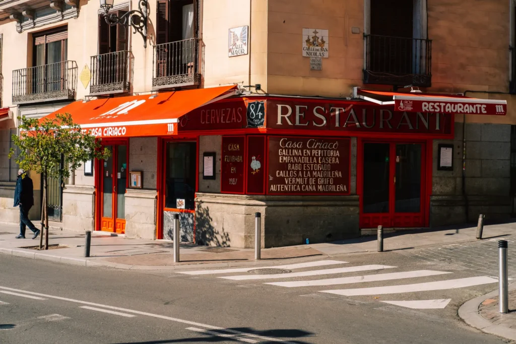 A classic corner restaurant, Casa Ciriaco, features bright red awnings and hand-painted text on its glass windows listing traditional Spanish dishes. In the foreground, a pedestrian walks along the sidewalk near a street crosswalk in front of the building.