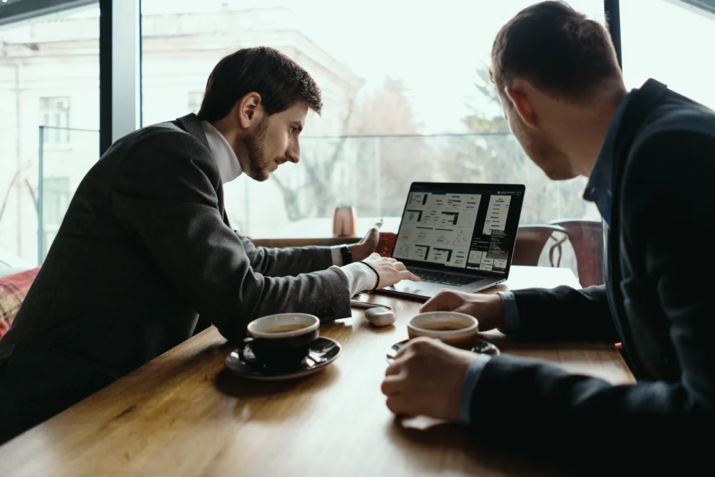 Two young businessmen sit across from each other at a wooden cafe table near a large window, sharing cups of coffee. One man types on an open laptop displaying design mockups while presenting the work to his colleague.