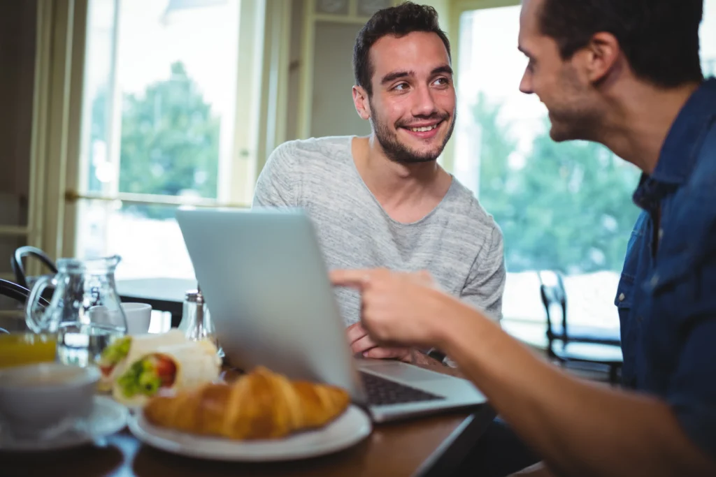 Two men sit at a cafe table with a open laptop, light food, and drinks while discussing content on the screen. One man points toward the screen while the other looks at him with a warm, friendly smile.