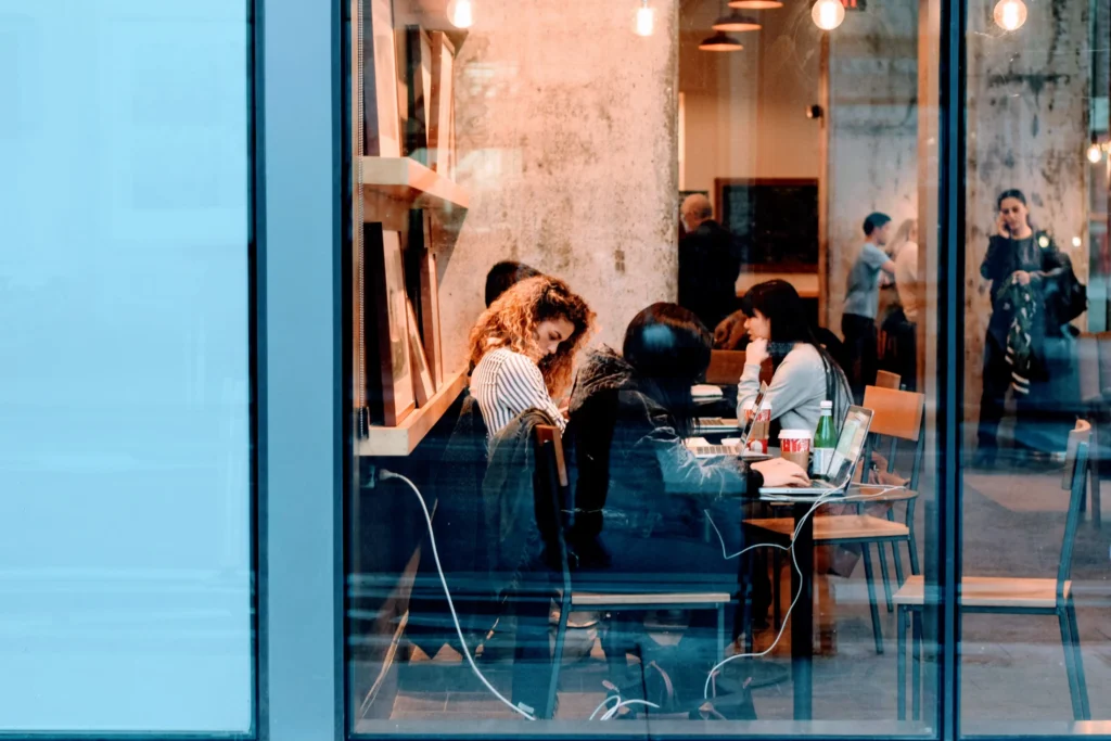 People sit inside a brightly lit coffee shop working on laptops and chatting, viewed through a large glass window pane from the outside street. The warm interior lighting contrasts with the cool blue reflections visible on the exterior glass facade.