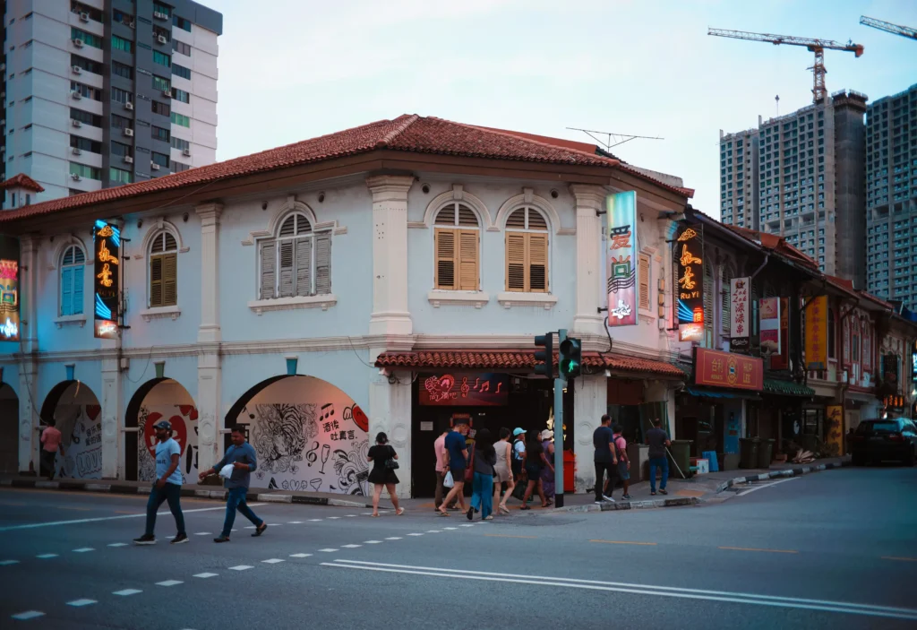 Pedestrians cross a street in front of a historic two-story shophouse adorned with glowing neon signs. Modern high-rise buildings and a construction crane tower in the background under a soft evening sky.