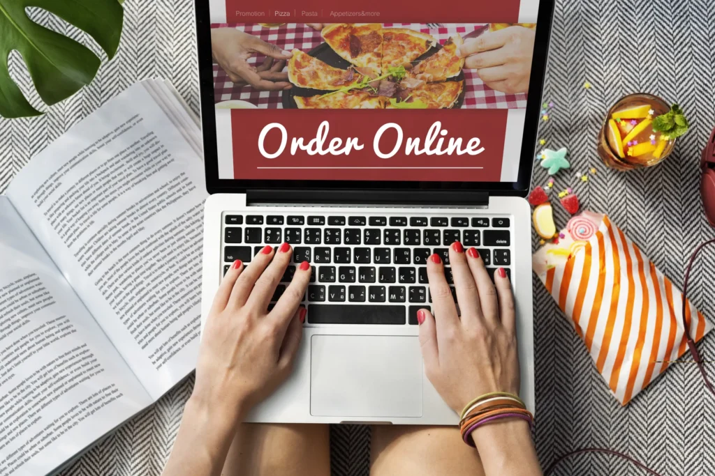 An overhead view shows a person typing on a laptop screen displaying a food delivery website with the text "Order Online" and an image of a sliced pizza. Surrounding the laptop on a patterned surface are an open book, a iced drink with fruit slices, and a colorful bag of snacks.