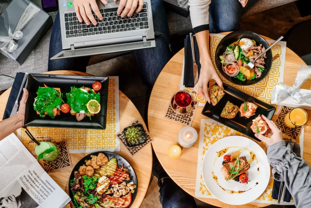 An overhead view shows people sharing a meal around round wooden tables filled with colorful dishes, salads, bruschetta, and drinks. One person works on a laptop at the top while others reach for food and enjoy the spread.