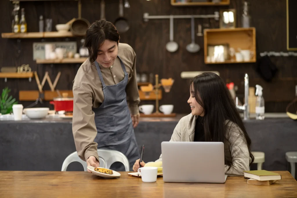 A server wearing an apron places a small plate with a pastry onto a wooden table for a customer. The woman sitting at the table smiles while working on her laptop with a mug and notepad nearby.