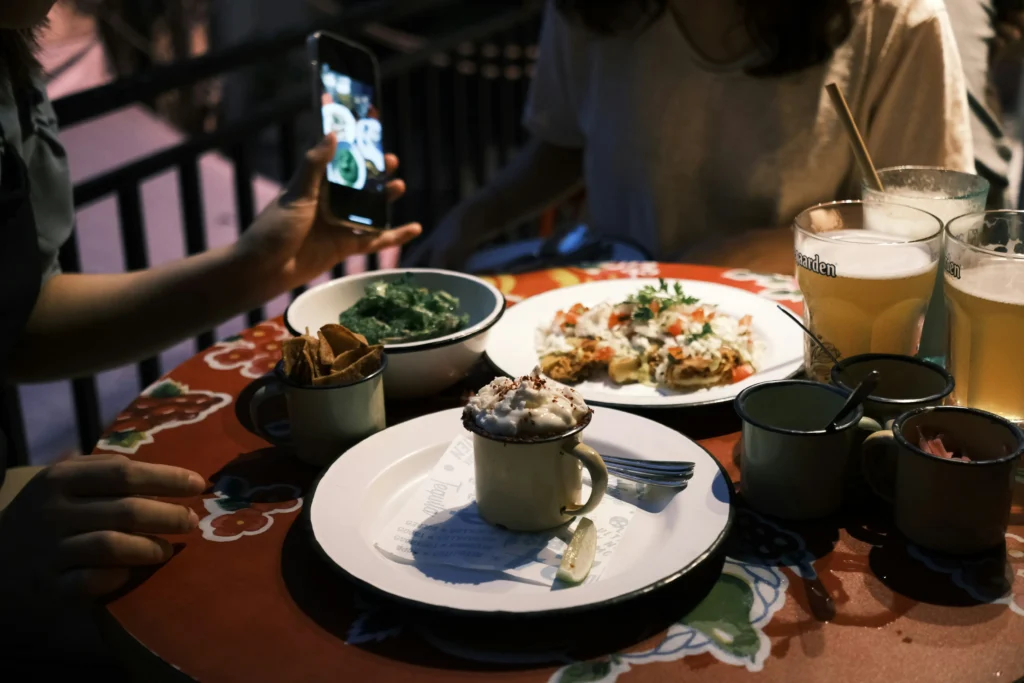 Two people sit at a patterned outdoor table filled with various dishes, enamel mugs, and glasses of beer. One person holds up a smartphone to take a photo of the spread, which includes a mug topped with whipped cream in the foreground.