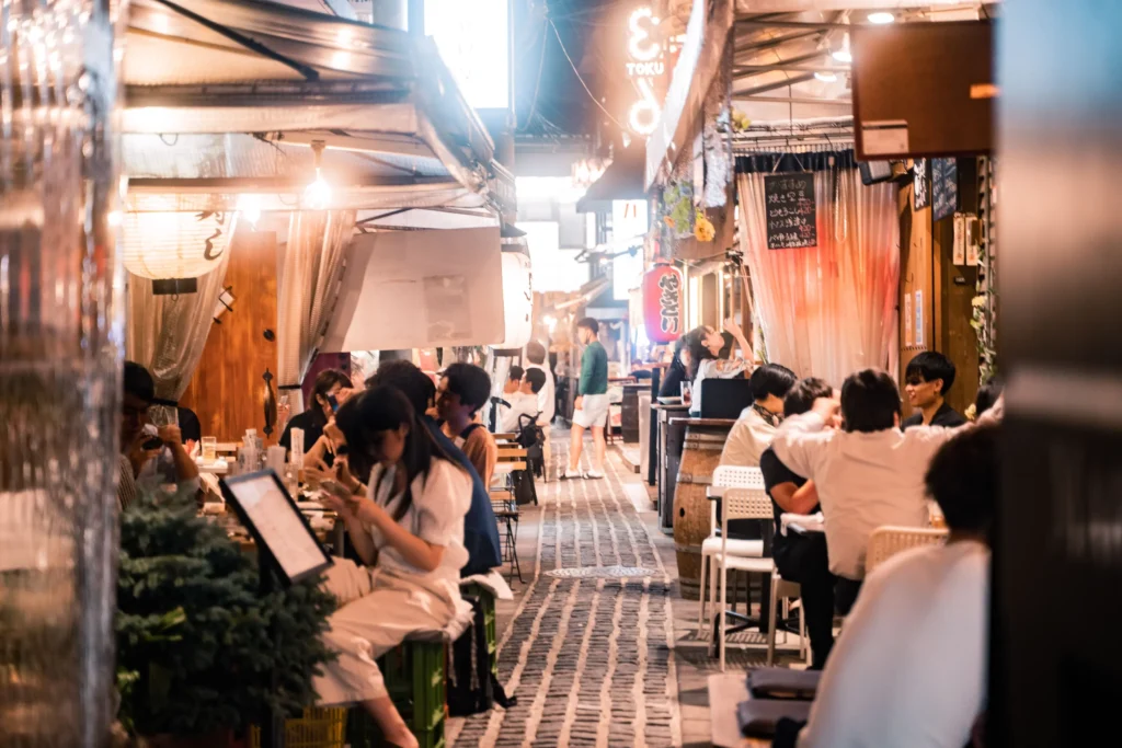 A bustling alleyway at night lined with open-air Japanese izakayas and outdoor food stalls. Patrons sit at small tables enjoying food and drinks under warm, glowing overhead lights and paper lanterns.