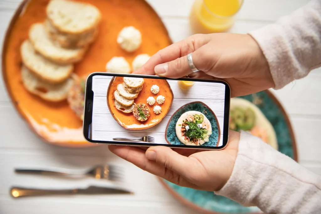 A person holds a smartphone in landscape orientation to capture a top-down photo of a colorful breakfast spread. The phone screen displays a clear photo of sliced bread, dips, and small dishes laid out on a white wooden table.
