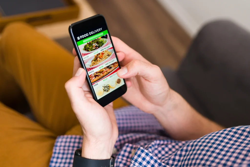 A person holds a smartphone while relaxing on a couch, browsing a food delivery app. The screen displays various menu items like salad, pasta, and pizza with "Add to Cart" options.